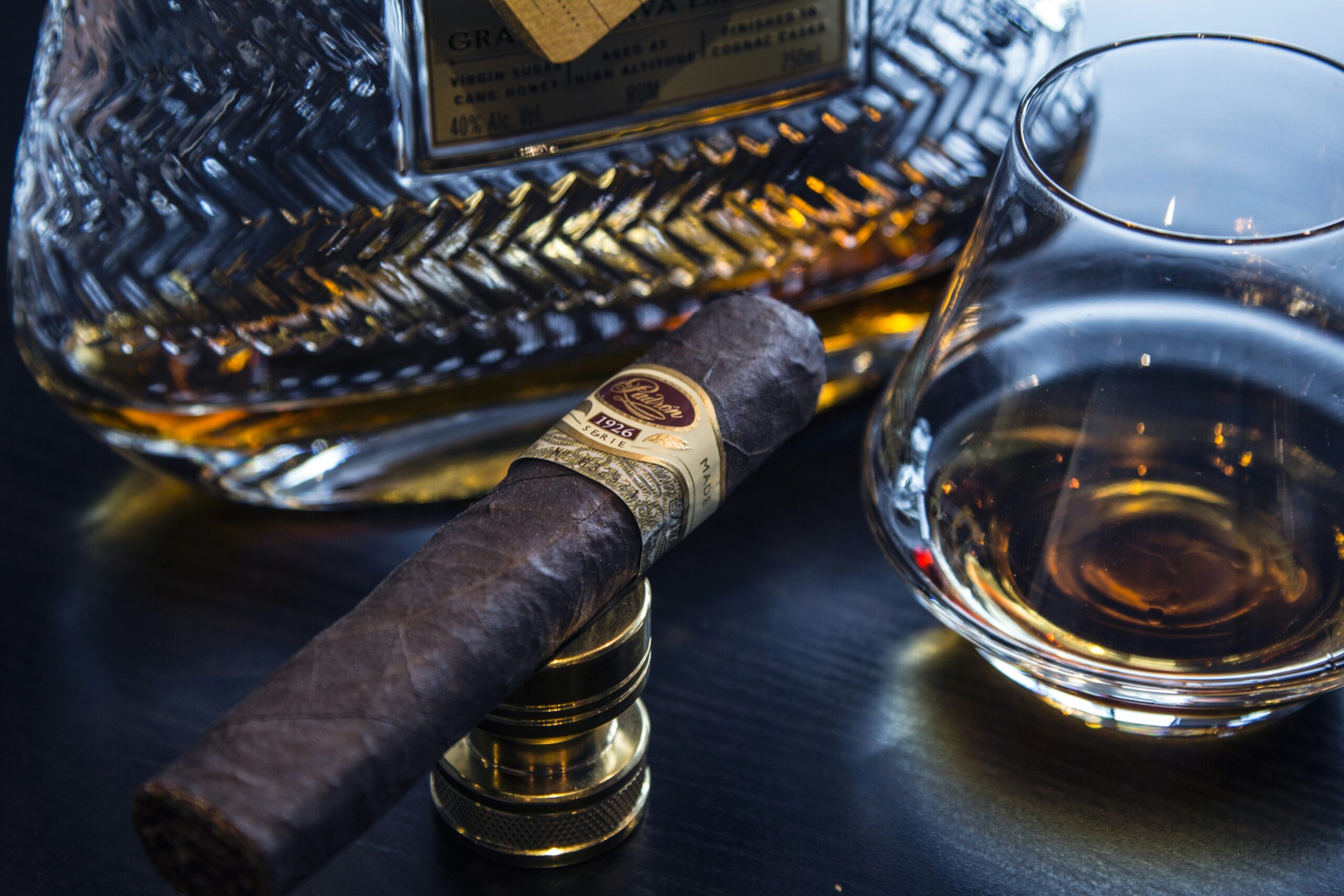 1200×675 px, warm editorial still life — a single cigar resting beside a wooden cutter and a lit match on a dark walnut surface, soft natural side light.